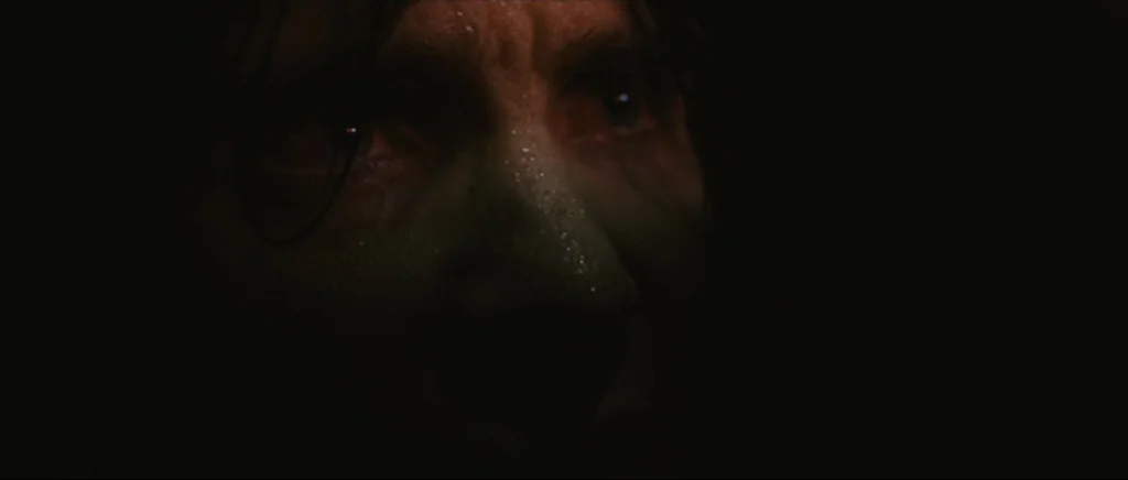 Sunshine movie still: closeup — A man's face emerges from total darkness in a dimly lit environment. Beads of sweat are visible on his skin as he…