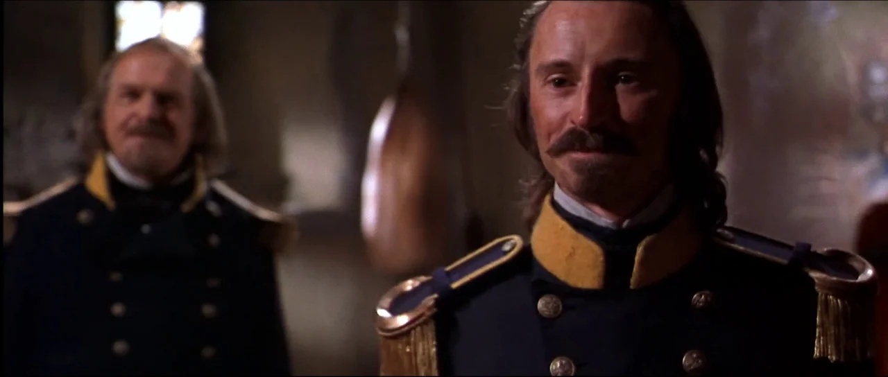 Ravenous (1999) movie still: medium — Two men in 19th-century military uniforms stand inside a dimly lit stone room. The men stand side-by-side…