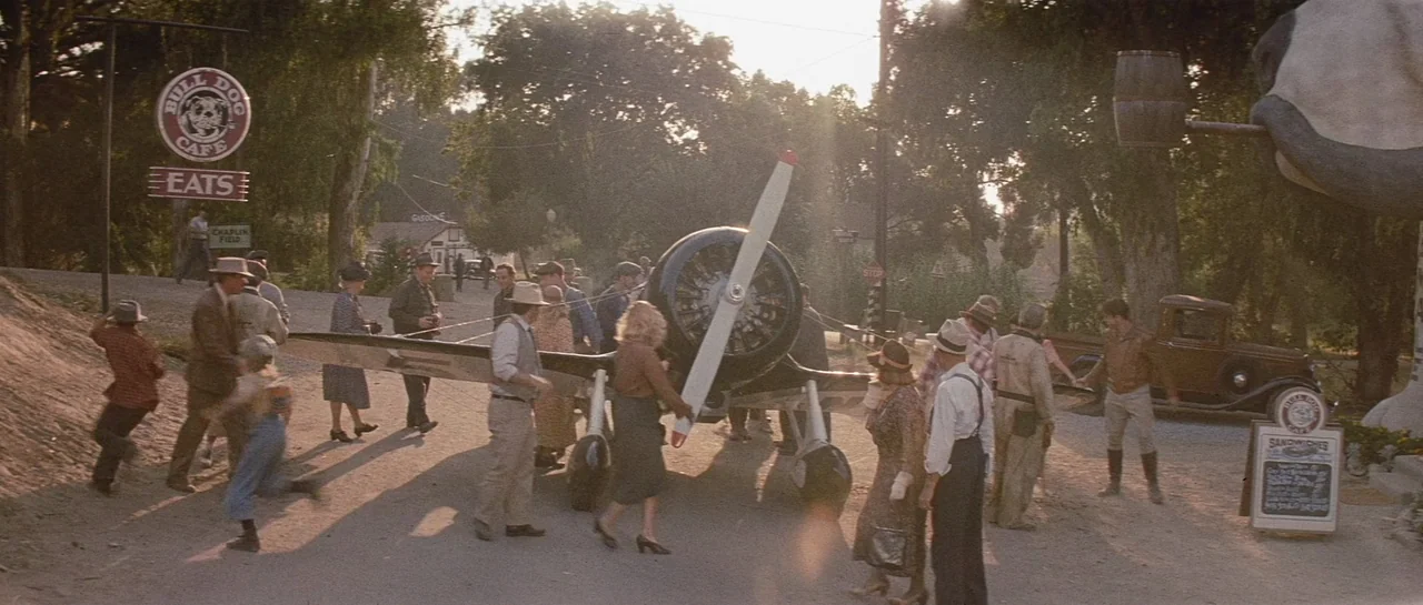The Rocketeer (1991) movie still: wide — A 1930s roadside cafe exterior features a small airplane parked on a dirt lot. A crowd of people in period…