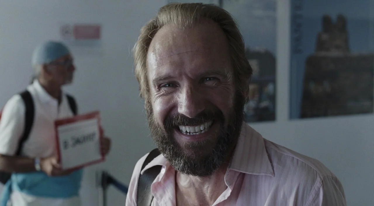 A Bigger Splash (2015) movie still: closeup — A man in a pink shirt smiles in an airport terminal hallway. A second person holding a sign waits out…