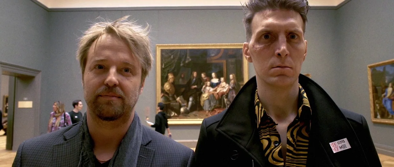 Depraved (2019) movie still: medium — A quiet museum gallery featuring large oil paintings displayed on cool grey walls. Two men stand in the…