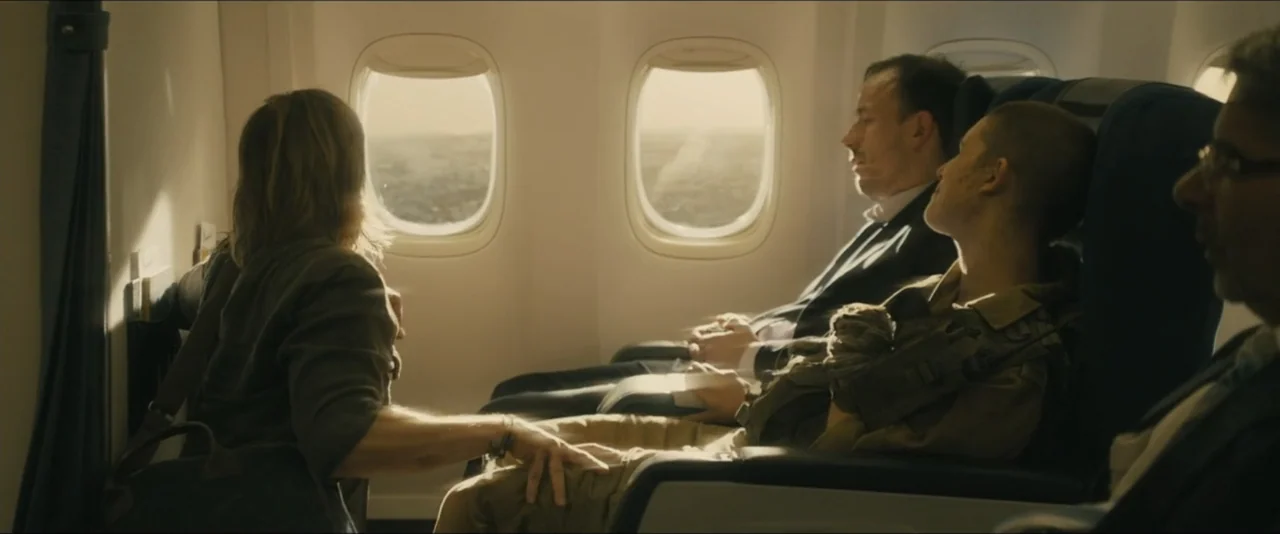 World War Z (2013) movie still: medium — Inside an airplane cabin with blue seats and bright light from two windows. A woman in a grey shirt reaches…