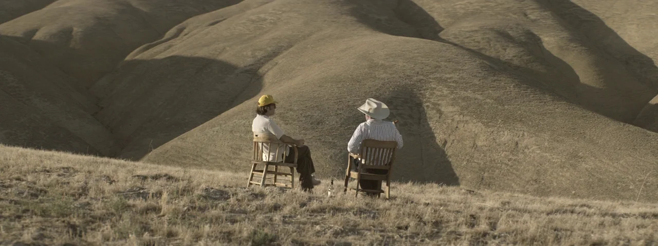 Entertainment (2015) movie still: wide — Two men sit in wooden chairs atop a parched, grassy mound under an open sky. The surrounding landscape…