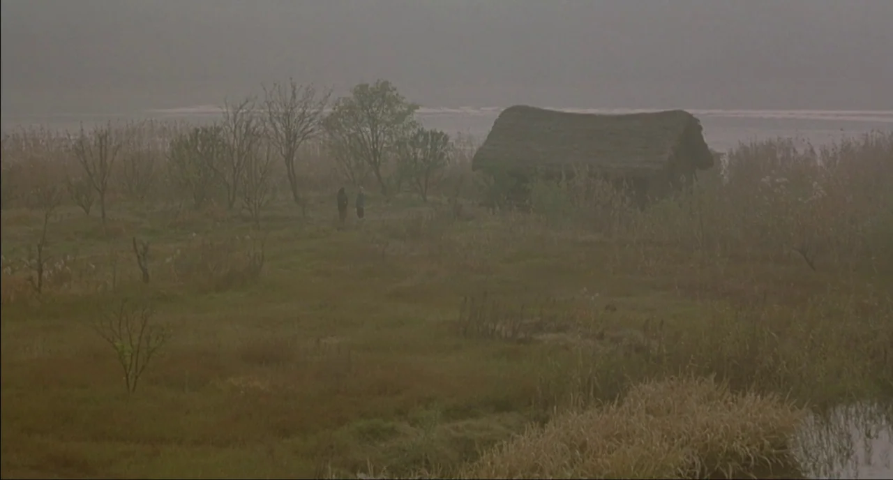 Shanghai Triad (1995) movie still: wide — A vast, misty marshland contains dry grass and several bare trees under heavy clouds. Two small figures…