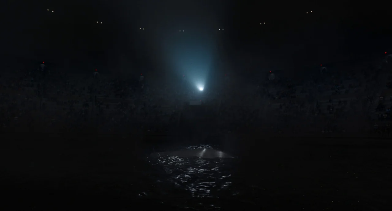 Beau Is Afraid (2023) movie still: wide — A dark, massive stadium interior with tiered seating filled with dim, indistinct silhouettes. Water covers…