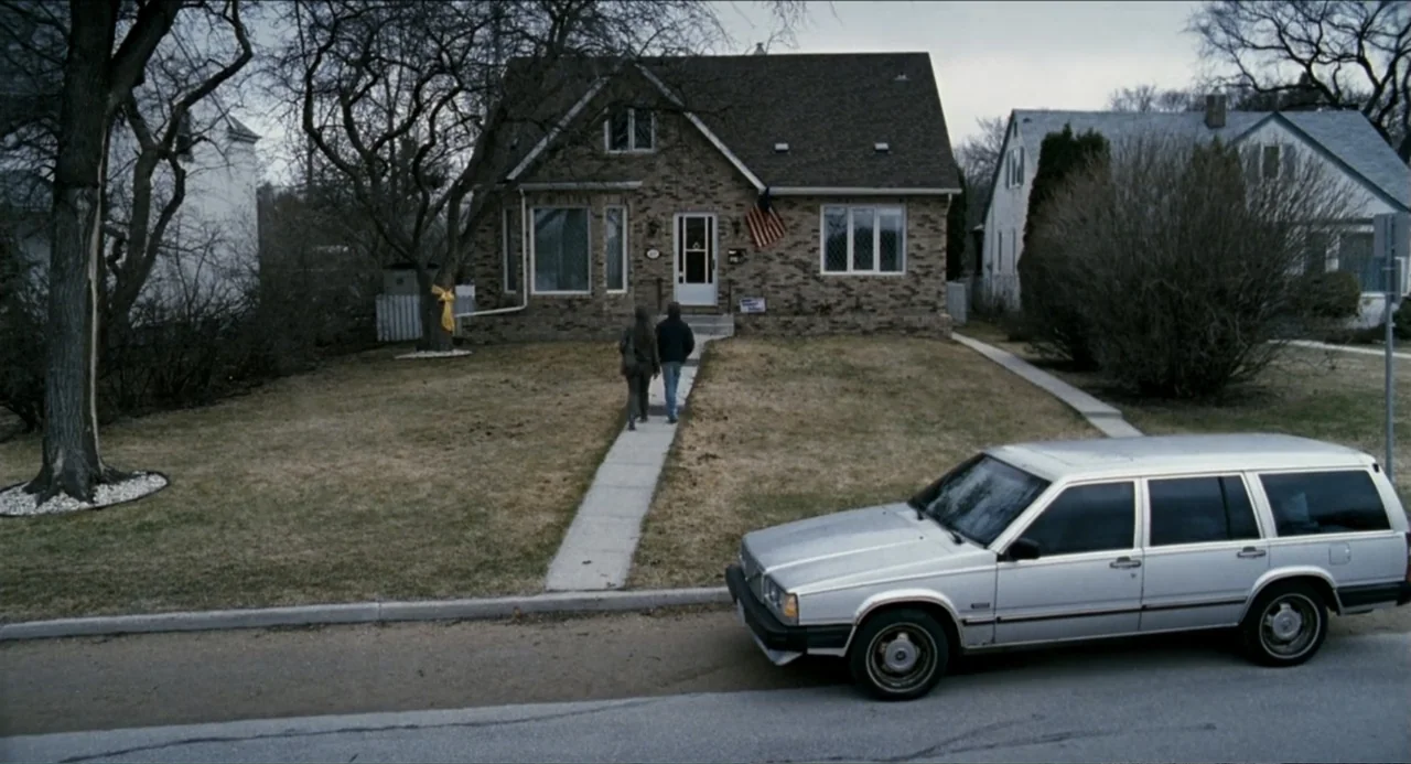 Colin Quinn: Red State, Blue State (2019) movie still: wide — A suburban brick house with a brown shingled roof stands behind a dry front lawn. Two…