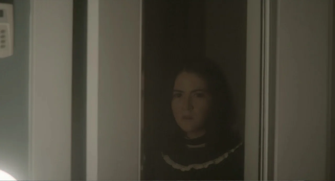 Orphan: First Kill (2022) movie still: closeup — A dark interior doorway frames a woman peering into a dimly lit space. She stands centered within…