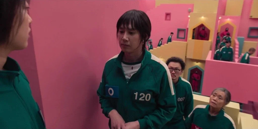 Squid Game (2024) movie still: medium — A pastel pink and yellow multi-level stairwell with geometric archways and stairs. Several people wearing…