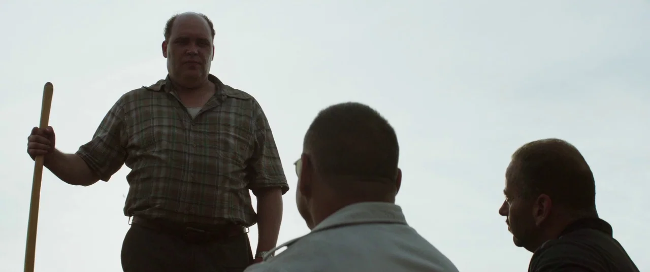 God’s Pocket (2014) movie still: medium — Three men stand against an overcast sky in an open, undefined outdoor space. A man on the left holds a…