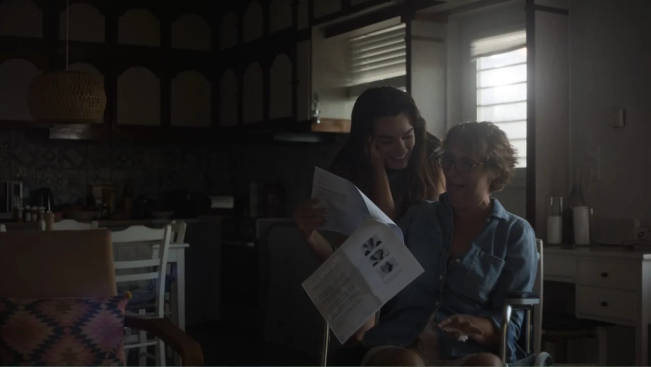 Hot Milk (2025) movie still: medium — A dimly lit room contains two women looking at documents near a window. A younger woman stands behind an older…