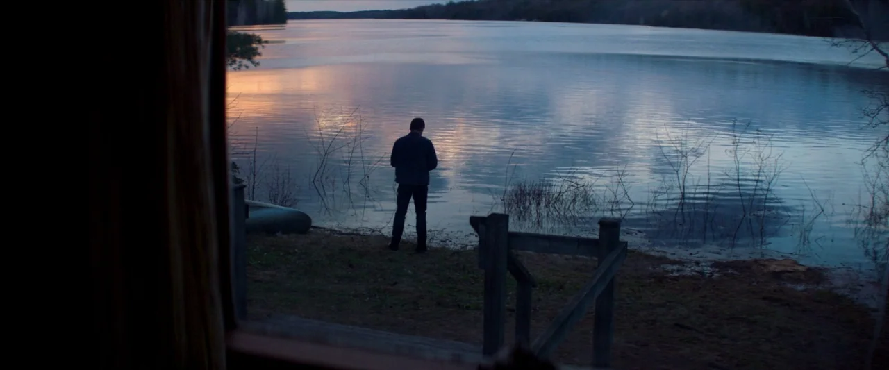 The Silencing (2020) movie still: wide — A man stands alone on the bank of a large lake at sunset. The wooden frame of a building opening partially…