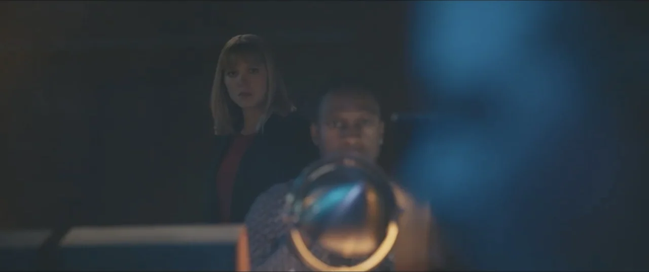 Zoe (2018) movie still: medium — A dark, dimly lit professional office environment during the night time. A woman stands behind a seated man as they…