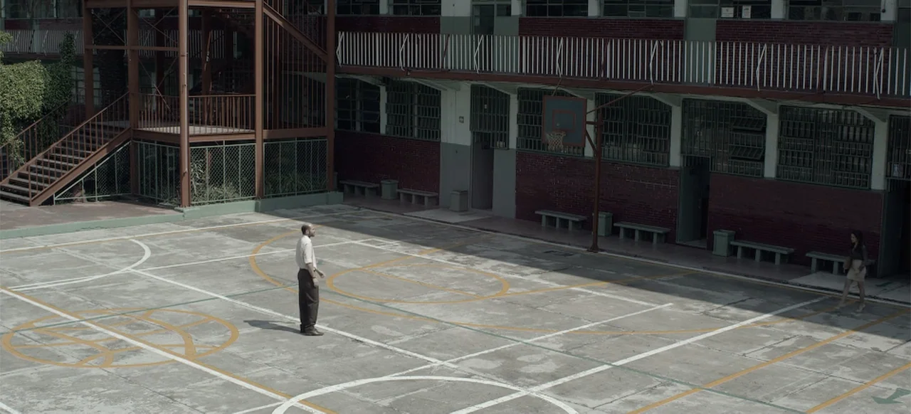 About the Deaths (2016) movie still: wide — An outdoor concrete sports area features weathered painted lines and dark brick-faced architecture.
