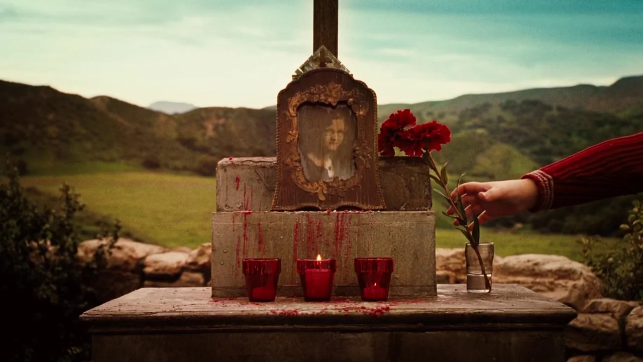 Nacho Libre (2006) movie still: medium — A stone memorial platform sits before rolling green hills under a sunset sky. A person's hand places a red…