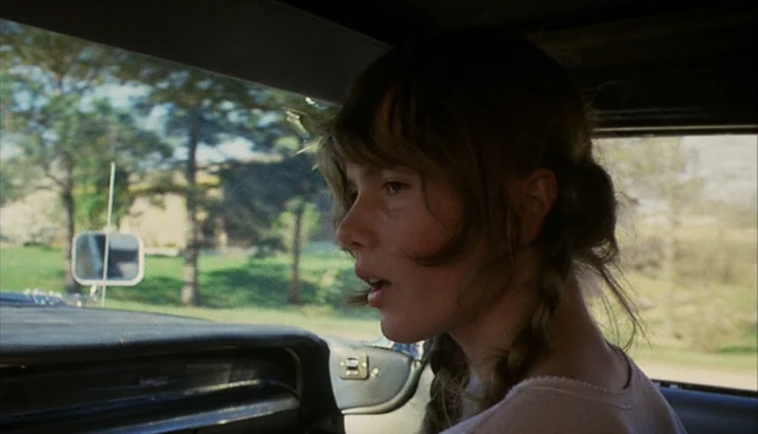 Cockfighter (1974) movie still: closeup — The interior of a moving car shows a green suburban landscape passing by outside. A teenage girl sits in…