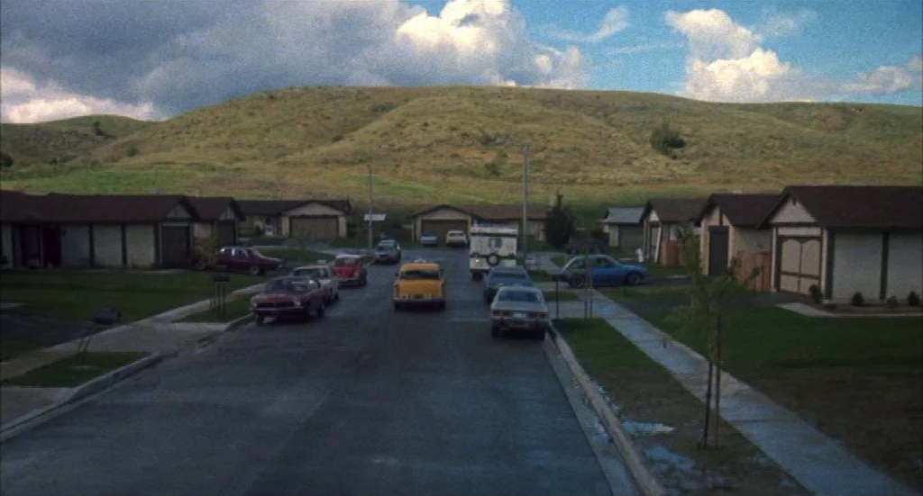 Melvin & Howard (1980) movie still: establishing — A quiet suburban neighborhood street lined with houses sits at the base of hills. Several…