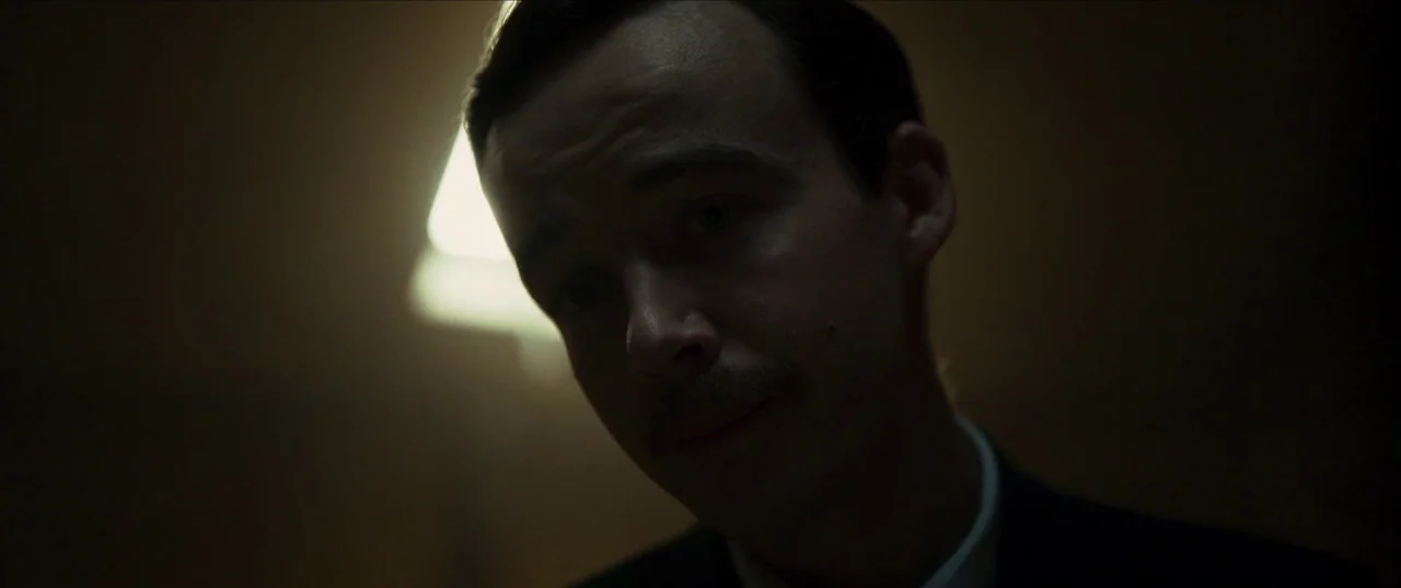 Earwig (2022) movie still: closeup — A dimly lit room serves as the setting for this close shot. A man in a suit looks down with a mustache visible…