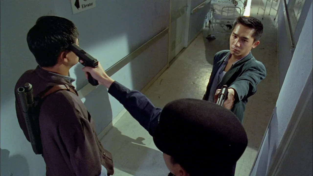 Hard Boiled (1992) movie still: medium — A dimly lit hospital corridor features a light blue wall with an elevator sign. Two men stand in a tense…