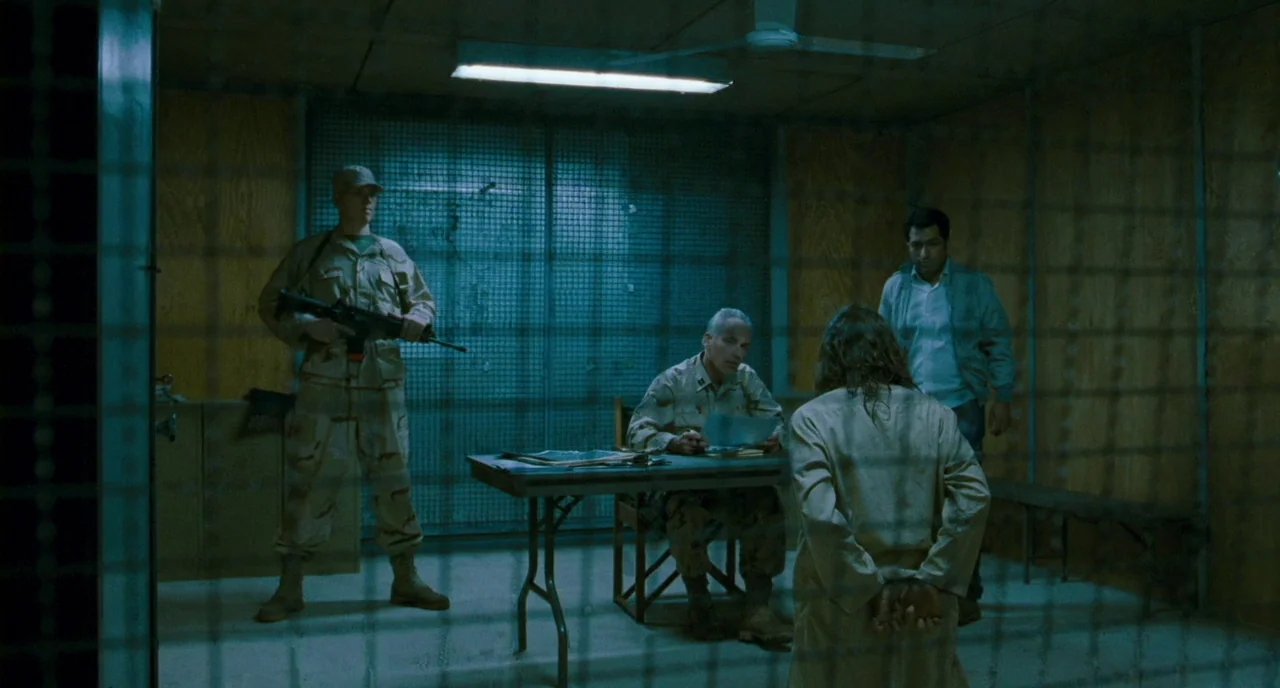 Essential Killing (2010) movie still: wide — A spartan room with plywood walls, metal grid fencing, and a folding table inside. Three men observe a…