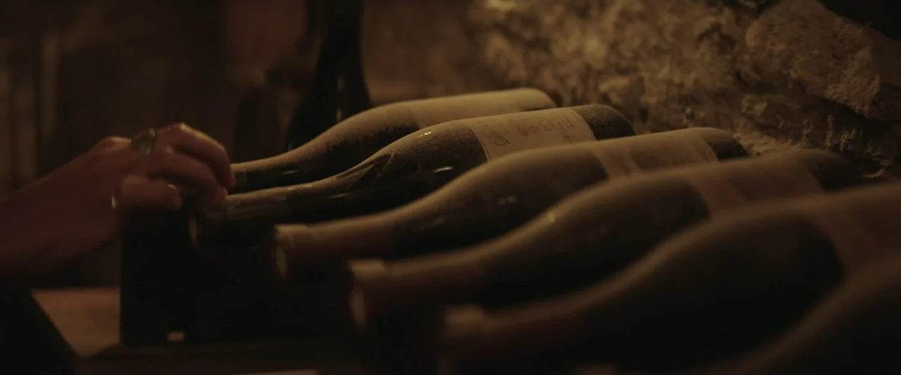 The Only One (2021) movie still: closeup — A dimly lit stone wine cellar contains shelves of dusty glass wine bottles. A human hand wearing a ring…