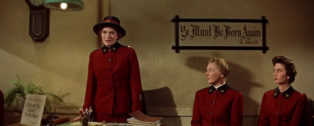 Guys and Dolls (1955) movie still: medium — A spartan room features beige walls adorned with a framed sign quoting scripture. Three women in…