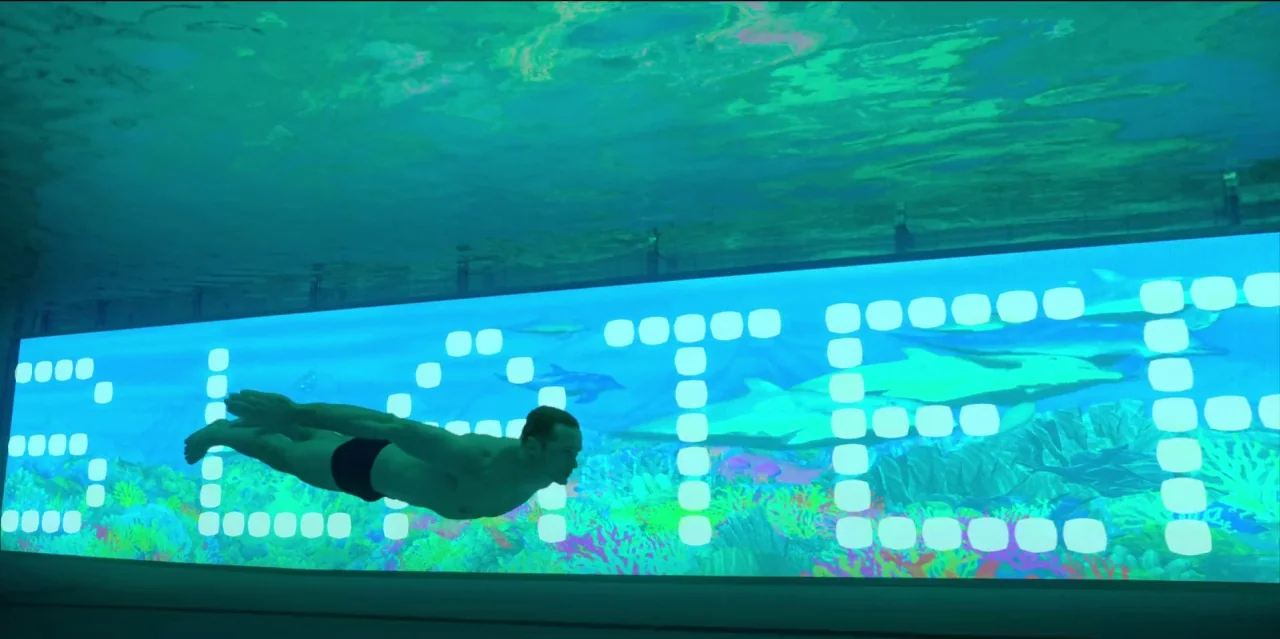 Mute (2018) movie still: wide — An athlete swims horizontally through an underwater environment illuminated by a massive digital screen. The screen…