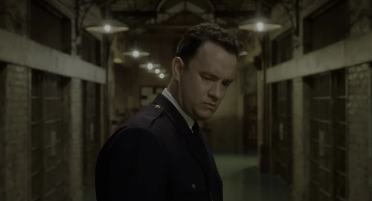 The Green Mile (1999) movie still: medium — A dark, narrow prison block corridor with brick walls and metal grated cells. A man in a dark uniform…