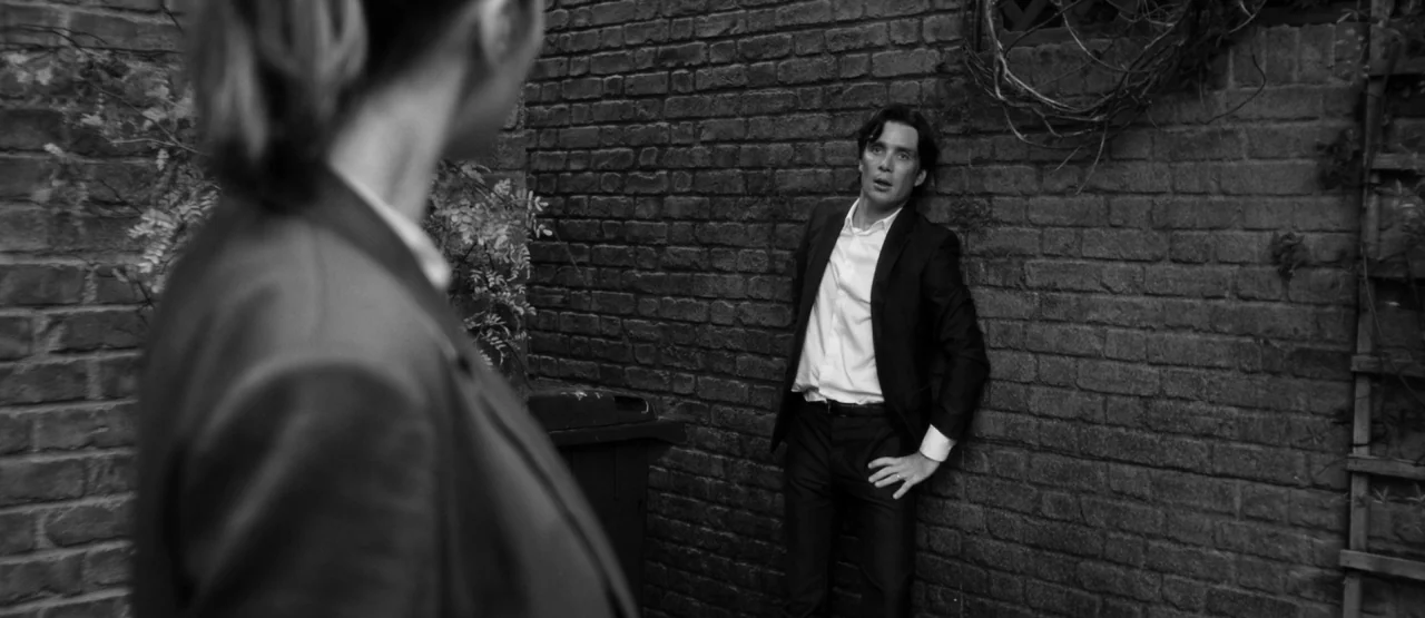 The Party (2017) movie still: medium — An outdoor area with a textured dark brick wall and climbing vines. A man in a suit stands against the wall…