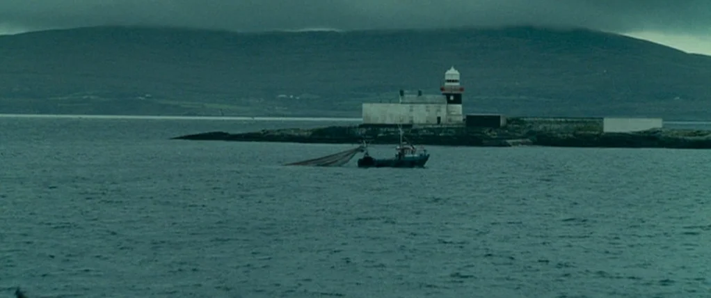 Ondine (2009) movie still: wide — A small fishing boat with a net operates on dark water near a rocky island. A lighthouse complex with white…