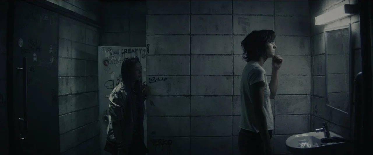 Kate (2021) movie still: medium — A dimly lit industrial restroom with concrete block walls covered in graffiti and stickers. One woman inspects her…