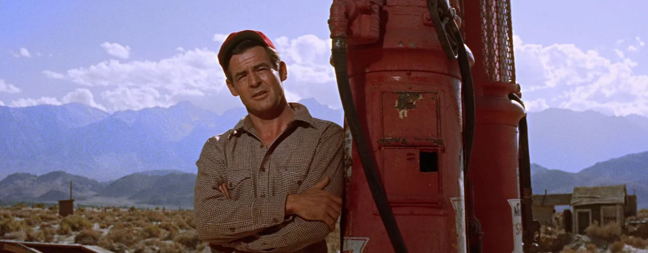 Bad Day At Black Rock (1955) movie still: medium — A rural setting features a vintage red gas pump under a clear sky. A man stands leaning against…