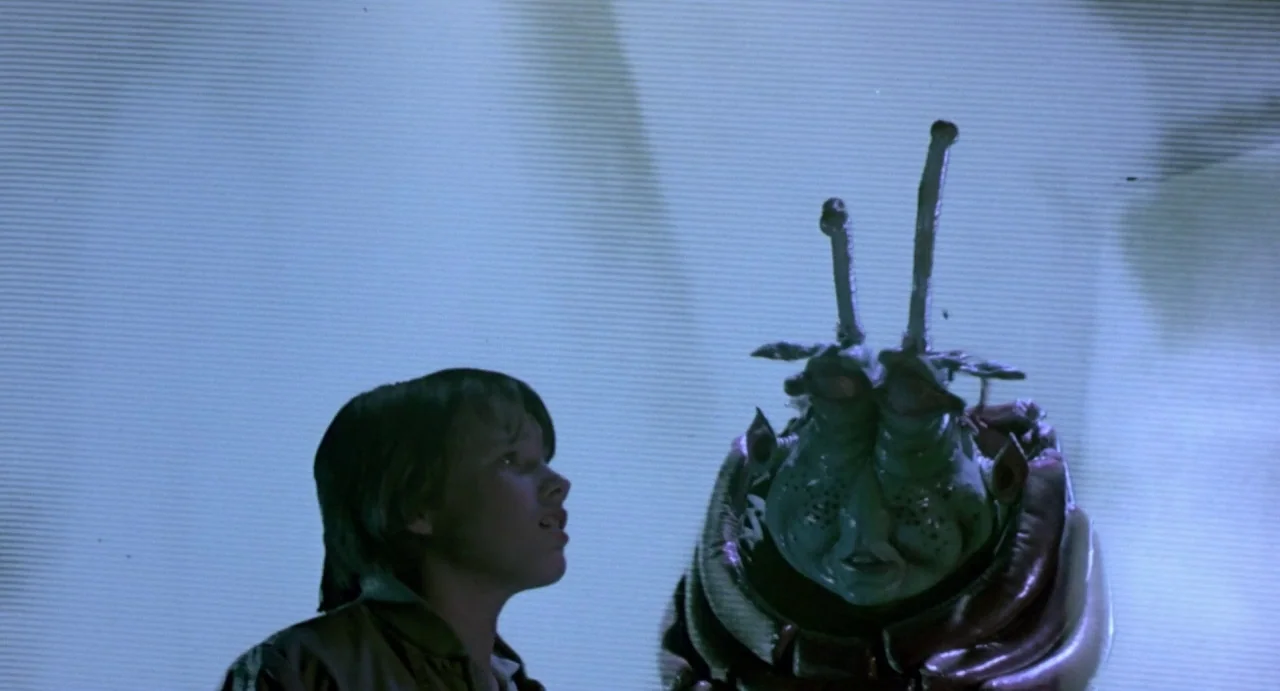 Explorers (1985) movie still: medium — A light-colored backdrop provides a flat, bright wall behind the two characters. A young boy stands next to a…