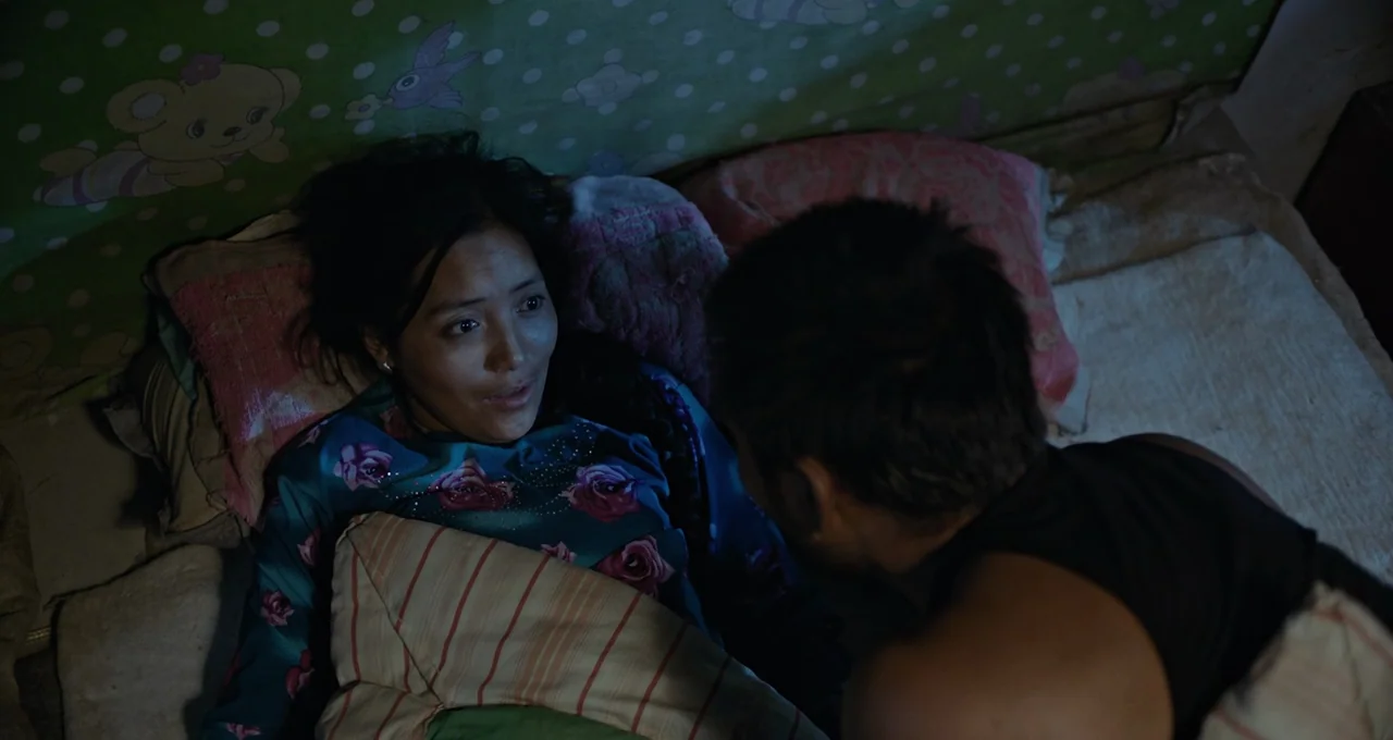 Balloon (2019) movie still: medium — A dark bedroom with a green patterned wall and pillows on a bed. A woman lies on the mattress while a person in…