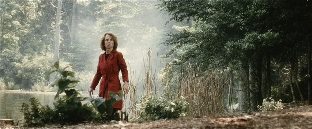 Minority Report movie still: fullbody — A woman stands on the bank of a lake surrounded by tall trees and brush. Wisps of light fog drift between…