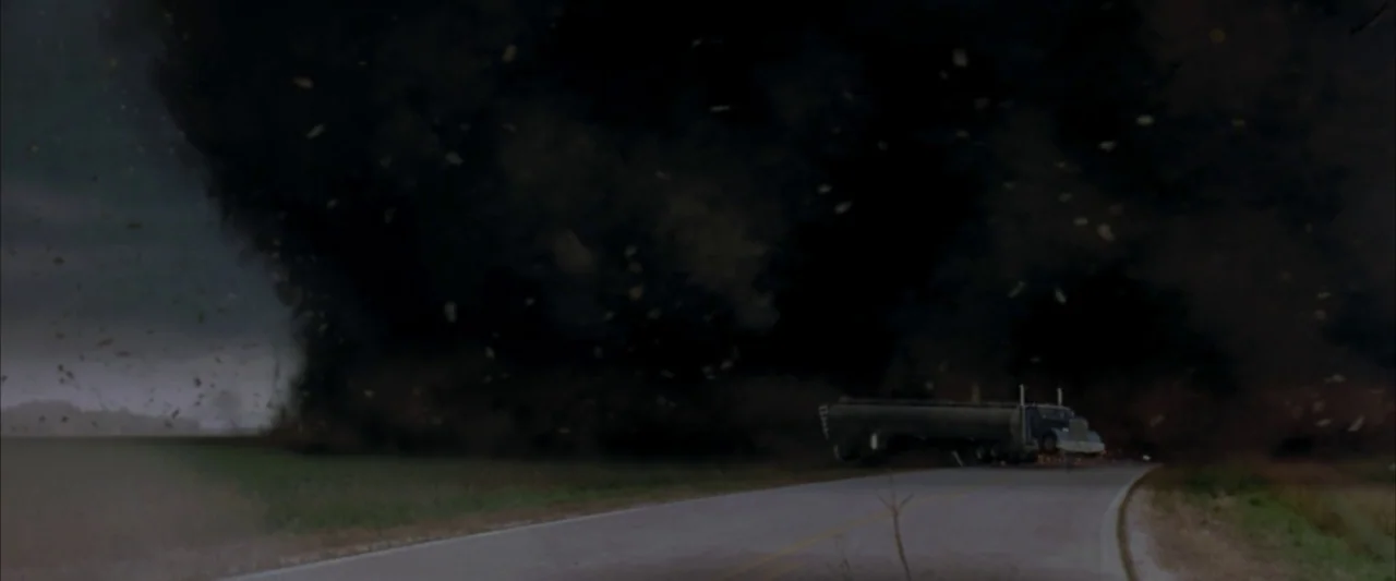 Twister (1996) movie still: wide — A dark, massive tornado churns across an asphalt road near an empty field. A semi-truck trailer sits on the road…
