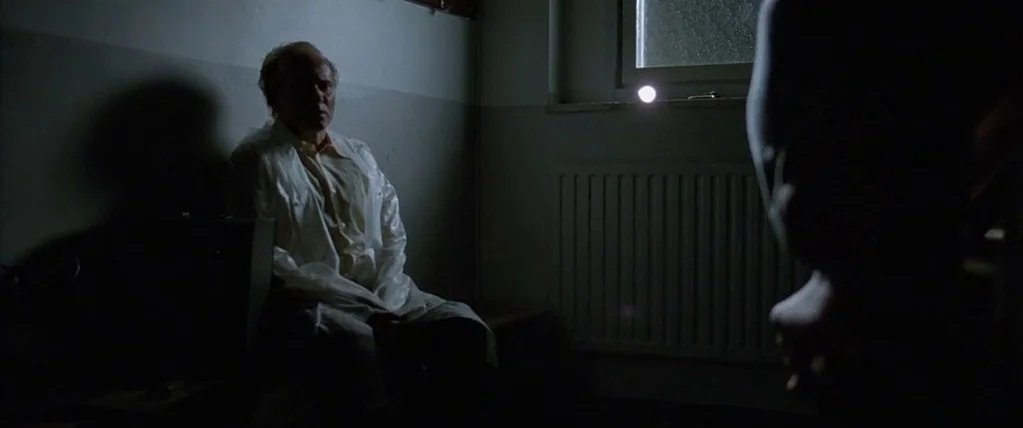 Let The Right One In (2008) movie still: medium — A dimly lit room with a radiator and a window at night. An elderly man in a white coat sits…