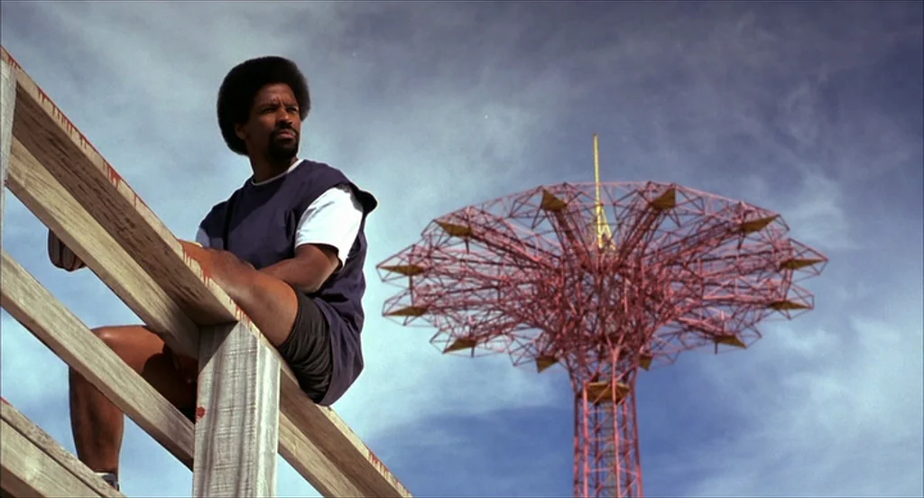 He Got Game (1998) movie still: medium — A man sits on a wooden railing against a backdrop of a clear blue sky. A tall, red, lattice-work amusement…