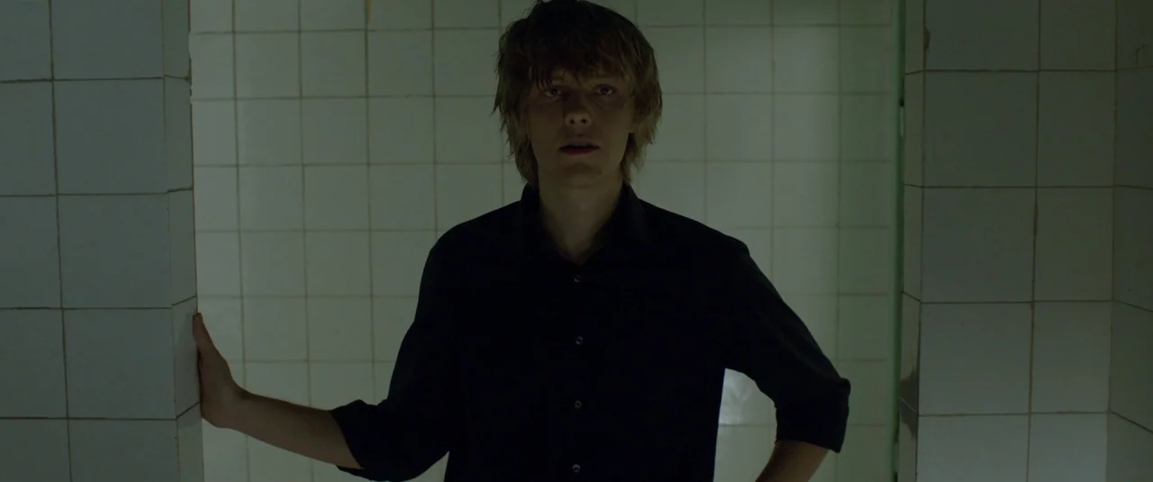 The Lure (2015) movie still: medium — A utilitarian restroom space is covered from floor to wall with small white tiles. A young man stands centered…