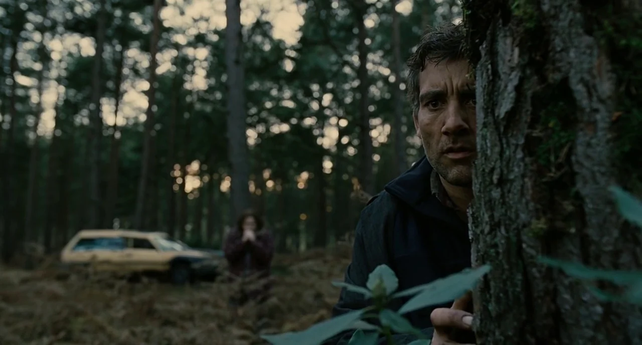 Children Of Men (2006) movie still: medium — A dense pine woods featuring a rough-barked tree in the foreground. A man watches from behind the tree…