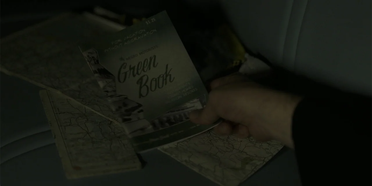 Green Book (2018) movie still: closeup — A hand holds a vintage travel guidebook over an unfolded paper map inside a vehicle. The scene is dimly lit…