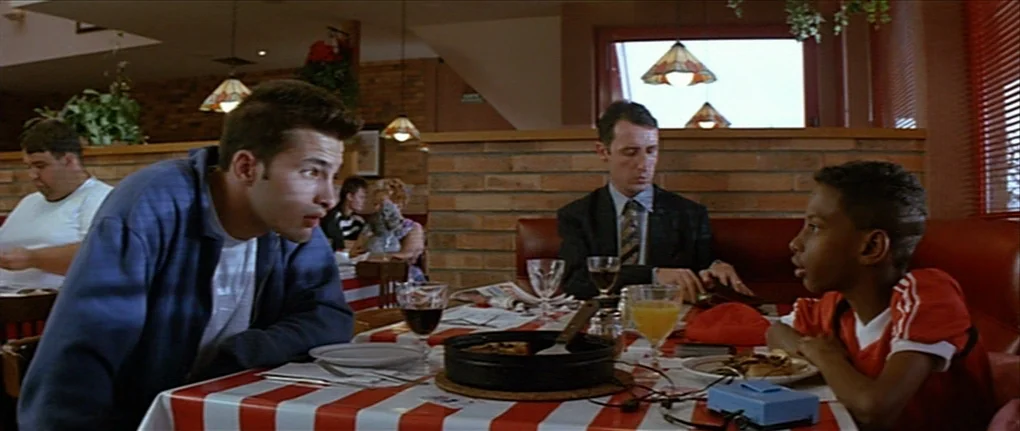 IP5 (1992) movie still: medium — A diner booth with red vinyl benches and a red and white checkered table cloth. A man leans forward to talk to a…
