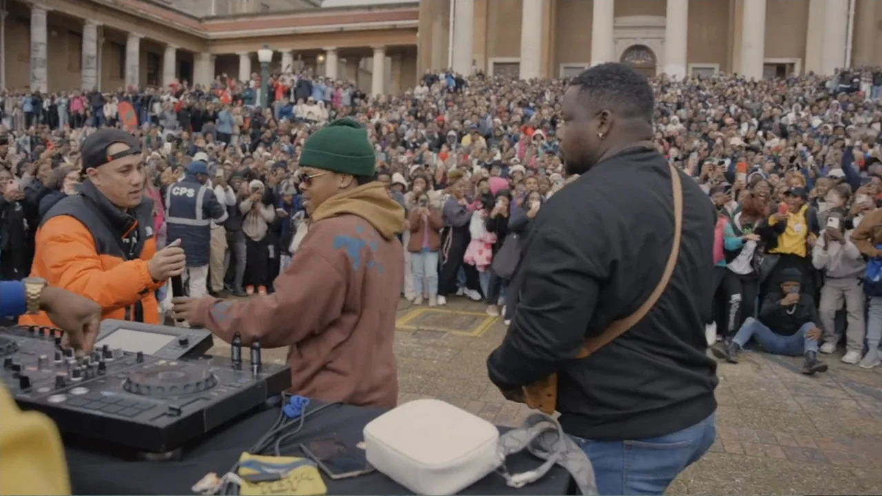 African Throne World Tour (2024) movie still: medium — A large outdoor plaza with a stone building facade serves as the backdrop. Three men stand…