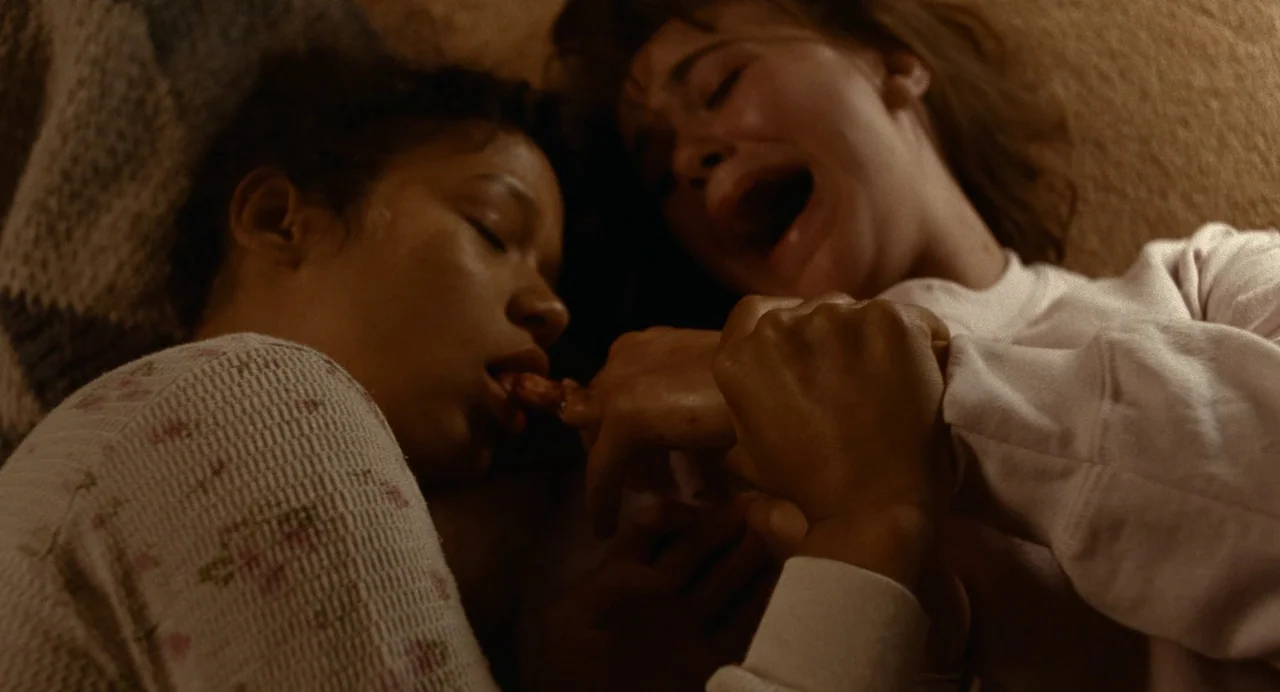 Bones and All (2022) movie still: closeup — Two women lie close together in a dimly lit, brown-toned indoor space. One person's hand is near the…