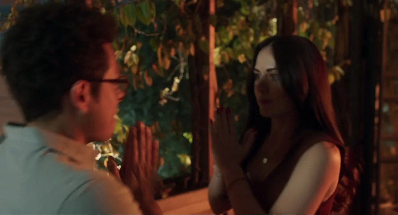 Casando a mi Ex (2023) movie still: medium — An interior space with a large glass window looking out at dark foliage. Two people stand facing each…