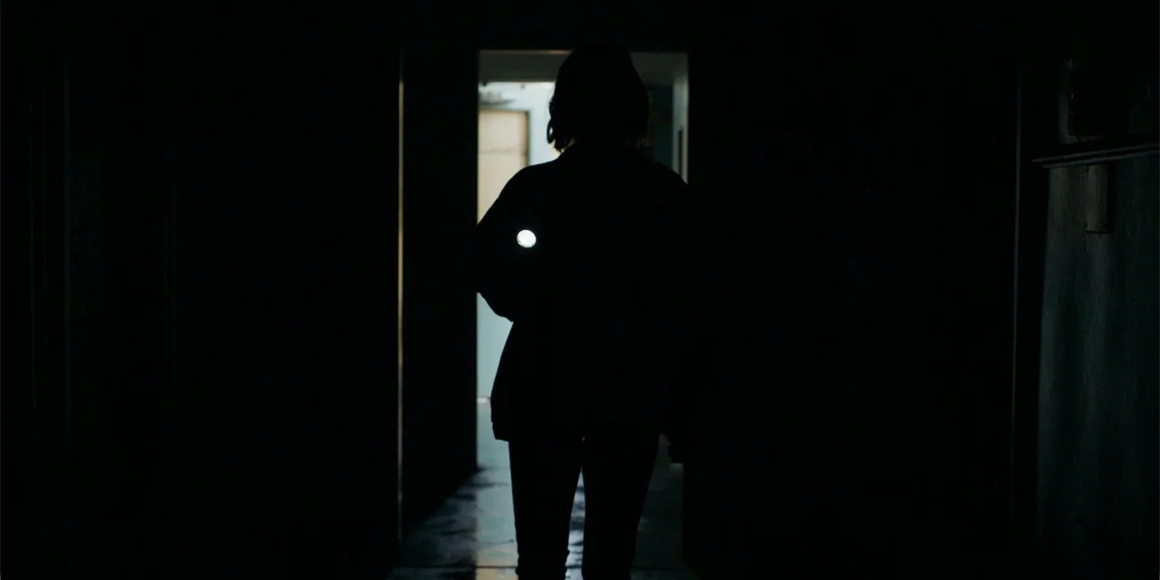 Demonic (2015) movie still: fullbody — A dark, empty corridor is illuminated only by a small, bright flashlight beam held by a person. The…