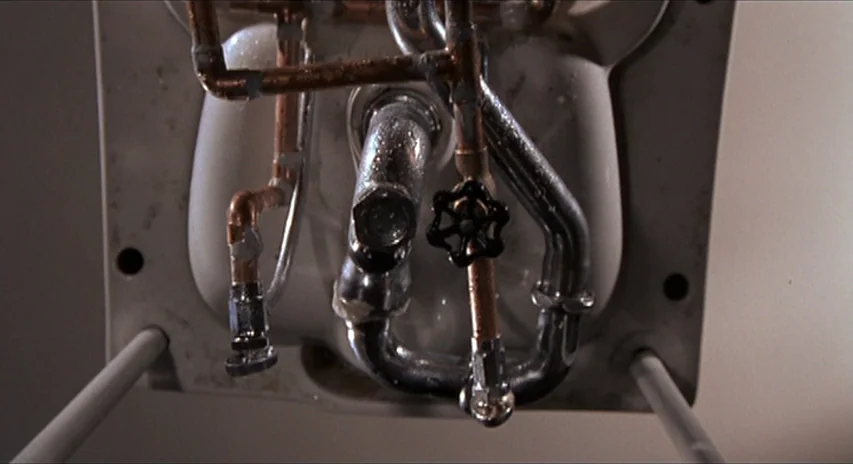 Blood Simple (1984) movie still: closeup — The underside of a bathroom sink shows exposed metal piping and a shutoff valve. Moisture droplets…