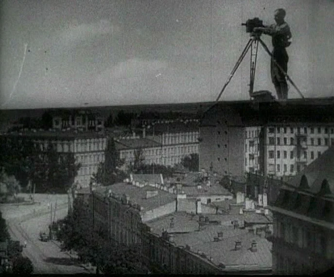 Man with a Movie Camera (1929) movie still: wide — A man operates a film camera on a tripod atop a city building roof. The surrounding landscape…
