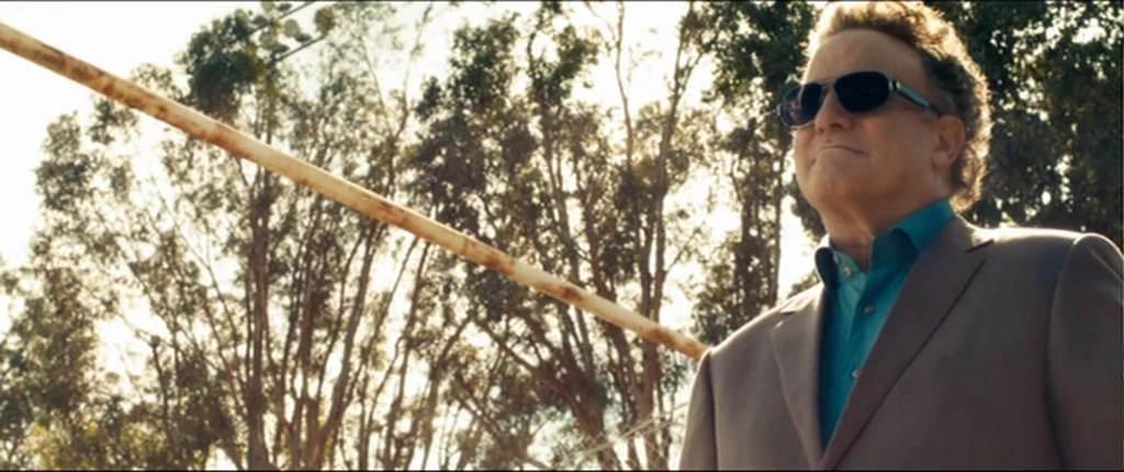 Drive (2011) movie still: medium — An older man stands outdoors in front of a line of tall thin trees. He looks up while wearing dark sunglasses…