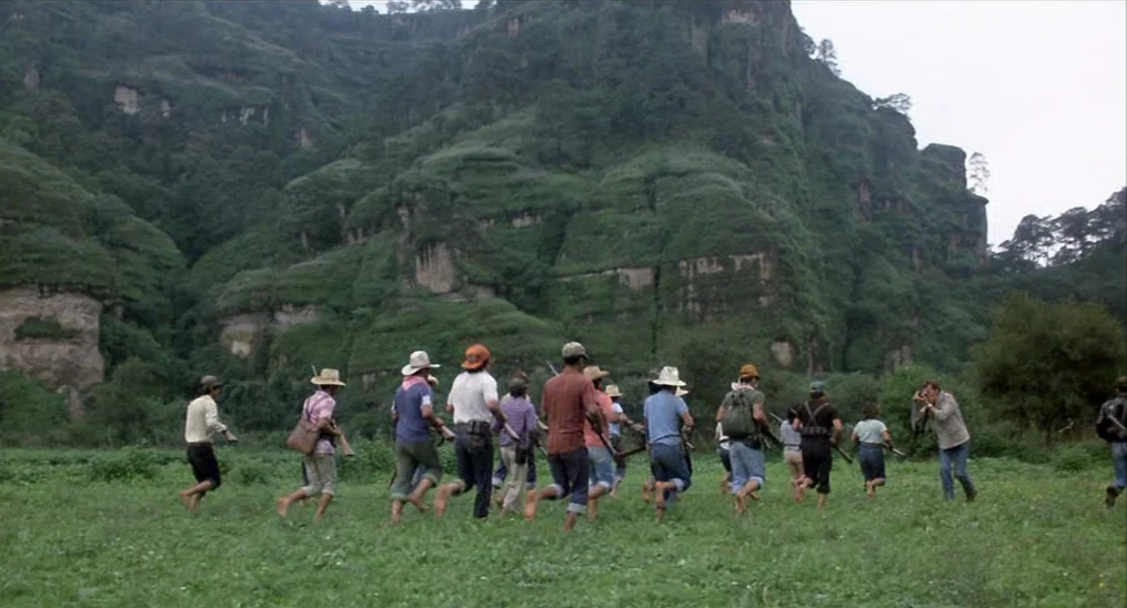 Salvador (1986) movie still: wide — A group of people walk through a large green field in front of mountains. The terrain features lush vegetation…