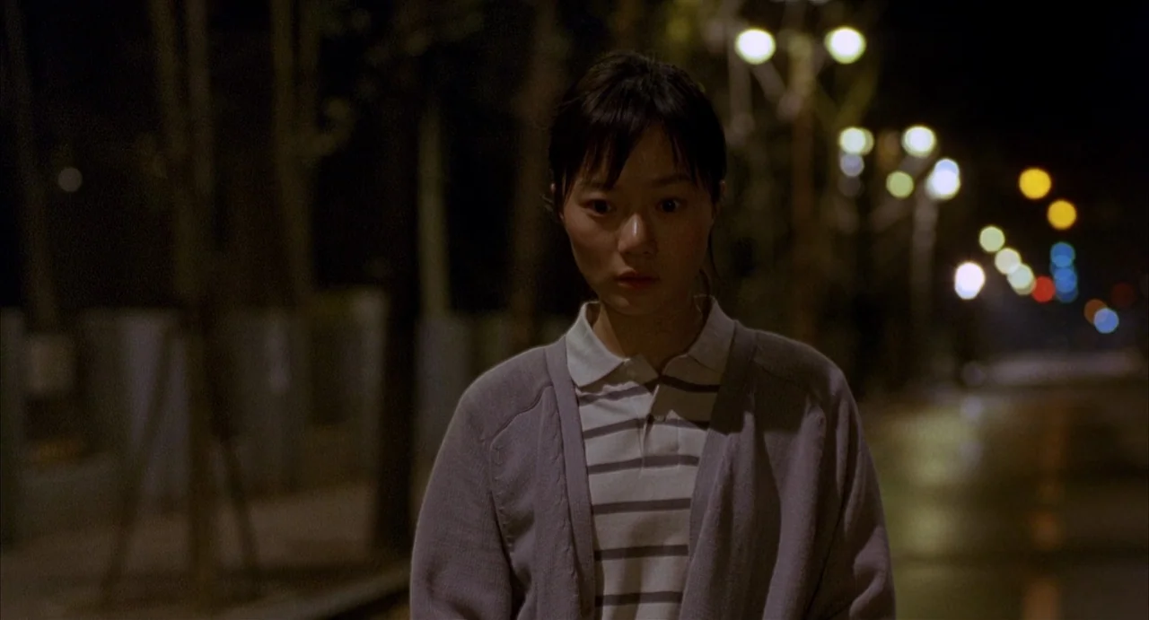 Barking Dogs Never Bite (2000) movie still: medium — A woman stands in the middle of a dark, empty paved area at night. Distant out-of-focus street…